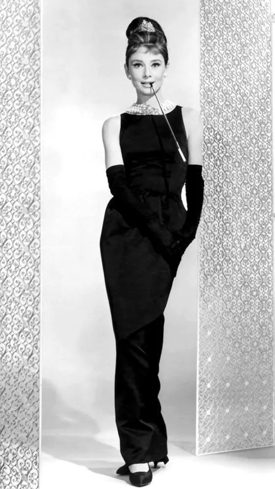 Audrey Hepburn de cuerpo entero con el vestido negro de Givenchy, guantes largos, collar de perlas, tiara y boquilla